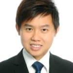 A young man with short dark hair, wearing a black suit, white shirt, and blue tie, smiles at the camera against a plain white background.