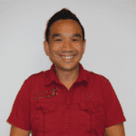 A person with short dark hair is smiling and wearing a bright red, short-sleeved button-up shirt. The background is plain and light-colored.