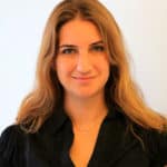 Une femme aux longs cheveux châtain clair, arborant un léger sourire et vêtue d'un chemisier noir, se tient debout devant un fond clair et uni.