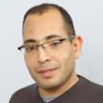 Un homme aux cheveux courts et foncés, portant des lunettes, est vêtu d'un pull foncé. Il fait face à la caméra et sourit légèrement devant un fond clair et uni.