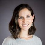 Une femme aux cheveux bruns mi-longs, vêtue d'un haut gris clair et portant des boucles d'oreilles pendantes, sourit à la caméra devant un fond uni gris foncé.