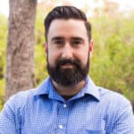 Ein Mann mit dunklem Haar und Vollbart, der ein blau kariertes Hemd trägt, steht draußen vor einem verschwommenen Hintergrund aus Bäumen.