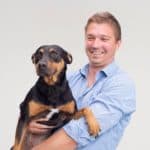 Vor einem schlichten hellen Hintergrund hält ein lächelnder Mann in einem blauen Hemd einen schwarz-braunen Hund im Arm.