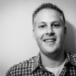 Portrait en noir et blanc d'un homme souriant aux cheveux courts et clairs, vêtu d'une chemise à carreaux par-dessus un t-shirt blanc, debout devant un fond uni de couleur claire.