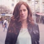 Eine Frau mit langen, welligen rotbraunen Haaren, die eine schwarze Lederjacke und ein helles Hemd trägt, steht draußen auf einer Straße in der Stadt, im Hintergrund sind verschwommene Geschäfte und Menschen zu sehen.