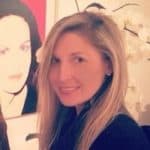 A woman with long blonde hair smiles at the camera. She is indoors, standing in front of a white orchid and a colorful portrait painting of a woman in the background.