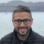 Ein Mann mit kurzen grauen Haaren, Brille und Bart lächelt in die Kamera. Er trägt eine dunkle Steppjacke und steht im Freien, im Hintergrund sind Wasser und eine Brücke verschwommen zu sehen.