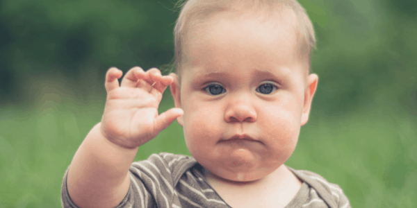 A baby in a grass field with his hand up waving goodbye.