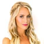 A woman with long, wavy blonde hair styled in loose curls and braided accents, wearing pink lipstick and a white top, poses against a white background.