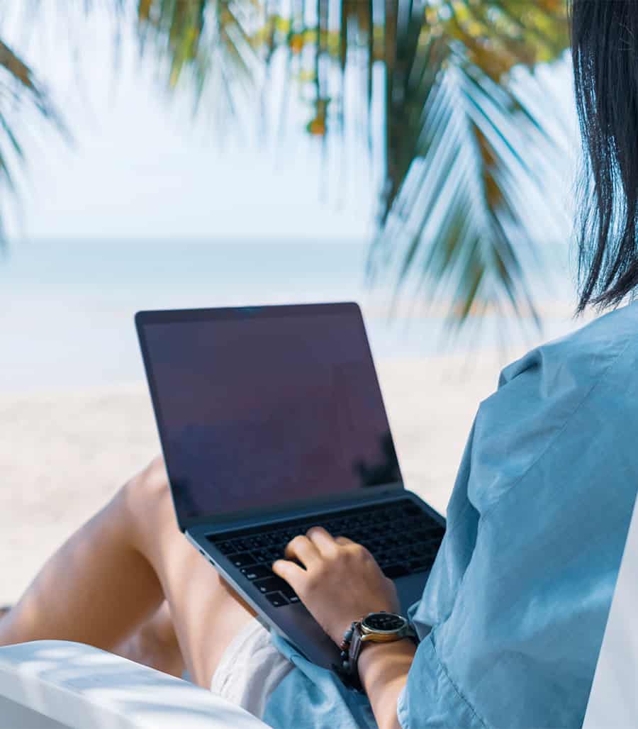 Woman lounging on with beach with palm trees while sitting on a chair on her laptop typing.