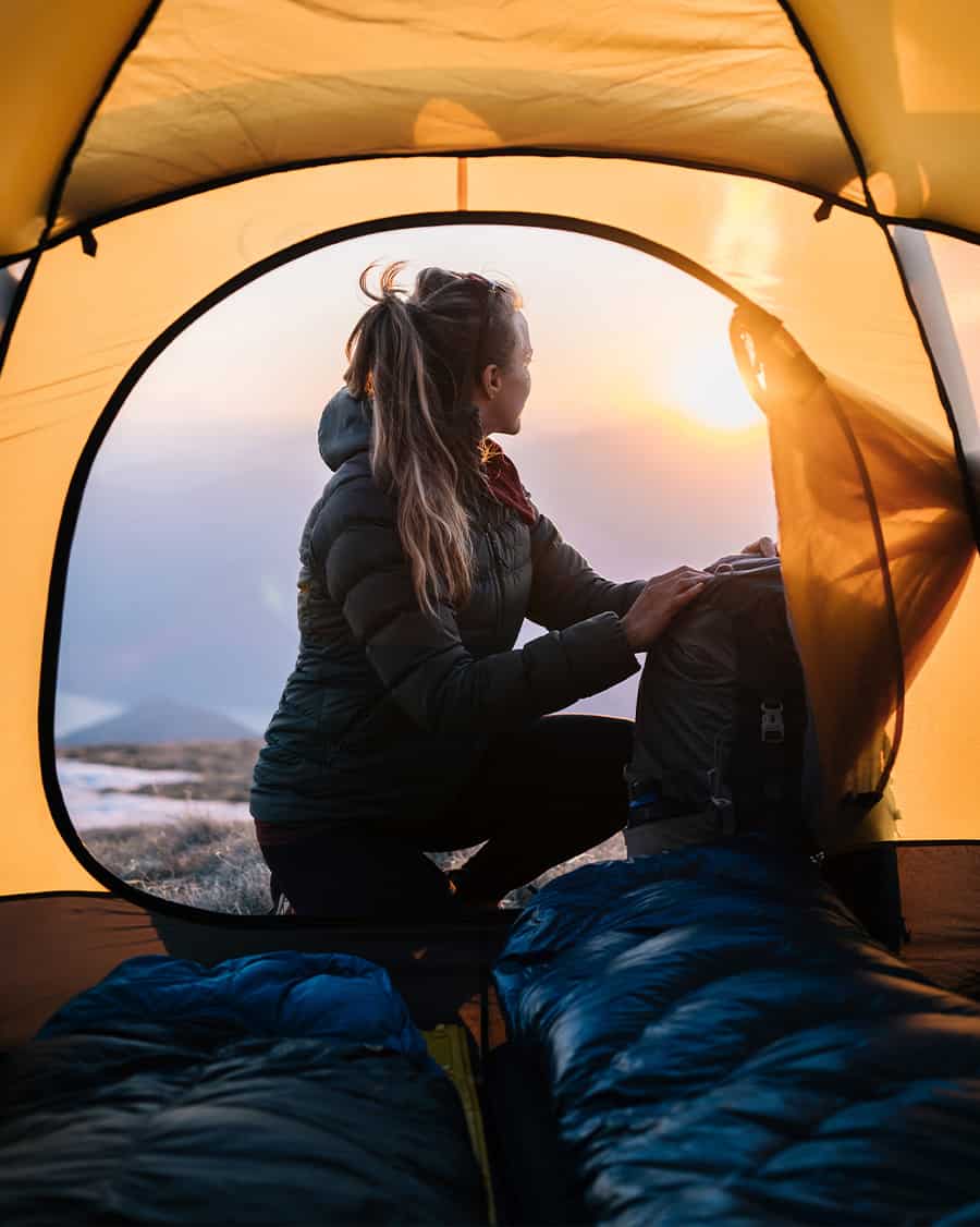Woman looking out of her camping tent at the sun setting in the elements.