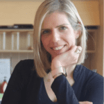 Une femme aux cheveux blonds et raides, vêtue d'un haut noir, sourit et pose son menton sur sa main. Elle se trouve à l'intérieur, avec des étagères et des classeurs en arrière-plan.