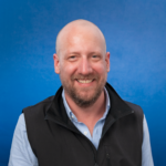 Ein lächelnder, glatzköpfiger Mann mit Bart, der ein hellblaues Hemd und eine schwarze Weste trägt, posiert vor einem einfarbigen blauen Hintergrund.