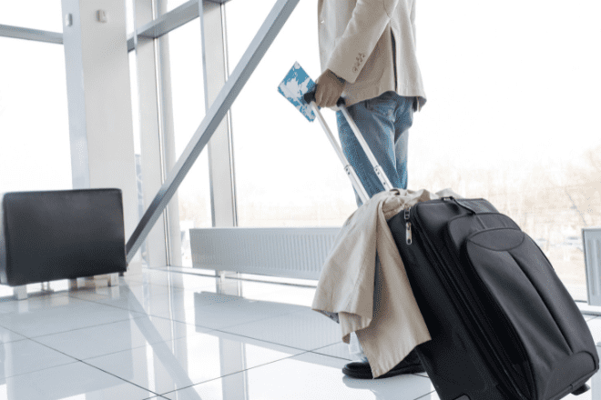 Une personne se tient dans un aéroport, une carte d'embarquement à la main, tirant une valise noire sur laquelle est posé un manteau beige, illustrant ainsi l'importance des expériences de voyage omnicanales qui relient toutes les étapes du parcours. Une lumière vive pénètre à travers les grandes baies vitrées en arrière-plan.
