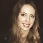 A young woman with long, light brown hair and blue eyes is smiling at the camera. She is wearing a dark top and a necklace. The background is dimly lit and out of focus.