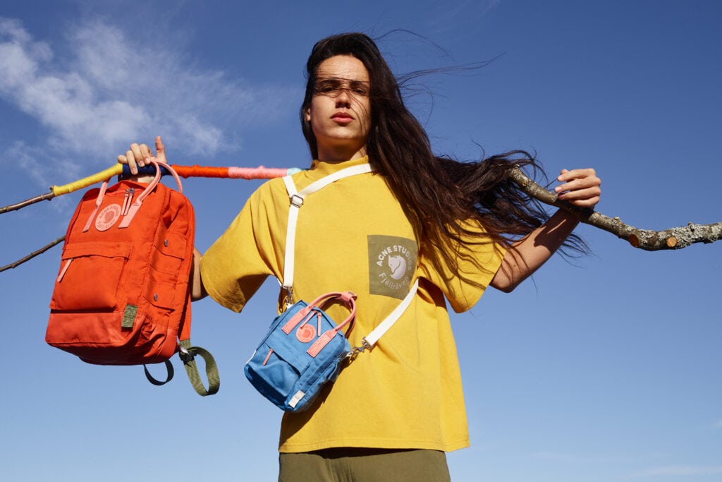 Advertisement for Fjallraven featuring a teenager in a yellow t-shirt holding a small blue backpack and a large red backpack against a blue sky backdrop.