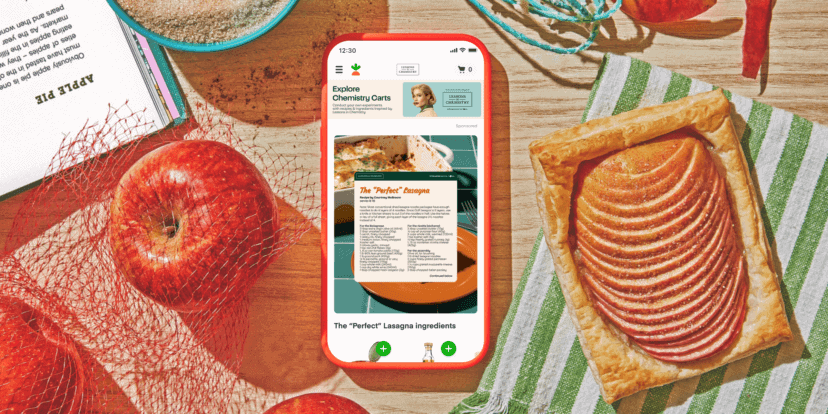 Ein Smartphone, auf dem ein Lasagne-Rezept angezeigt wird, liegt auf einem Tisch, auf dem Äpfel, ein aufgeschlagenes Buch, ein roter Netzbeutel und eine rechteckige Apfeltorte auf einem blauen Tischtuch liegen – eine einladende Szene, die ohne Weiteres Teil einer Instacart-Werbekampagne sein könnte, die die Freude am Kochen zu Hause feiert.
