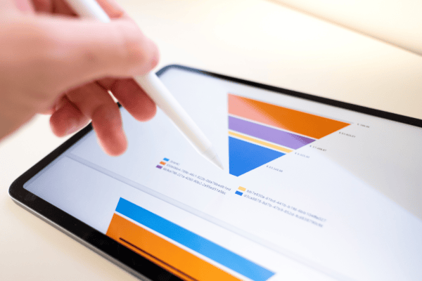 Eine Hand, die einen Stylus hält, zeigt auf den Bildschirm eines Tablets, auf dem bunte Diagramme zu sehen sind, darunter ein Dreiecksdiagramm und ein Balkendiagramm, was auf eine Datenanalyse oder -präsentation hindeutet.