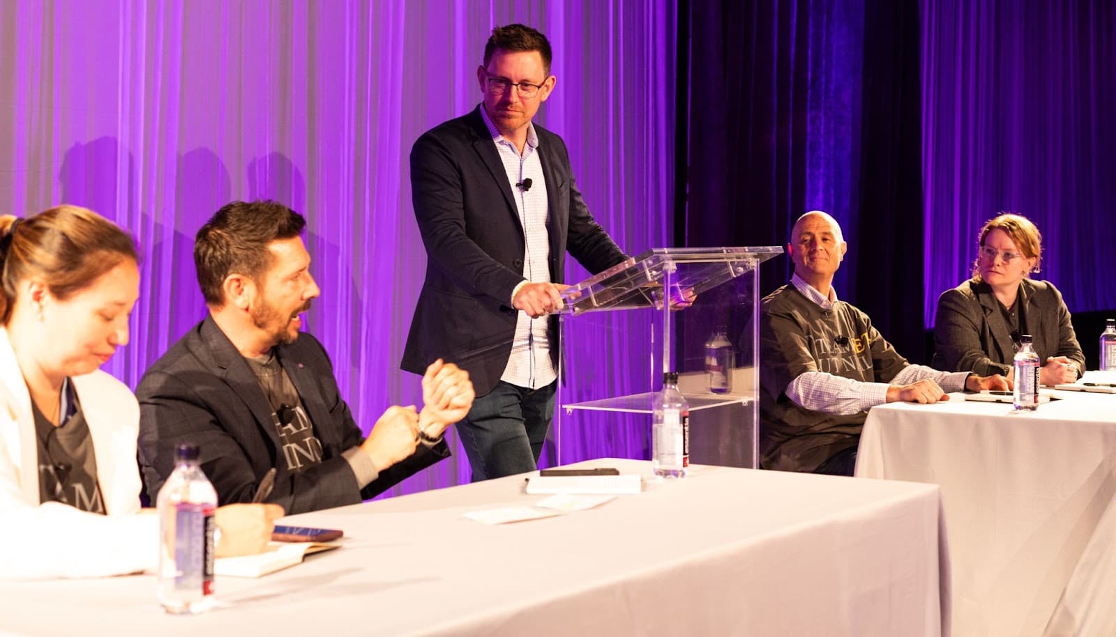 Four people sit at two tables facing a man standing at a clear podium. The background is purple-lit curtains. Bottles of water and papers are on the tables, suggesting a focused atmosphere where the group appears engaged in a discussion or debate—perhaps exploring whether retail media is retail or media.
