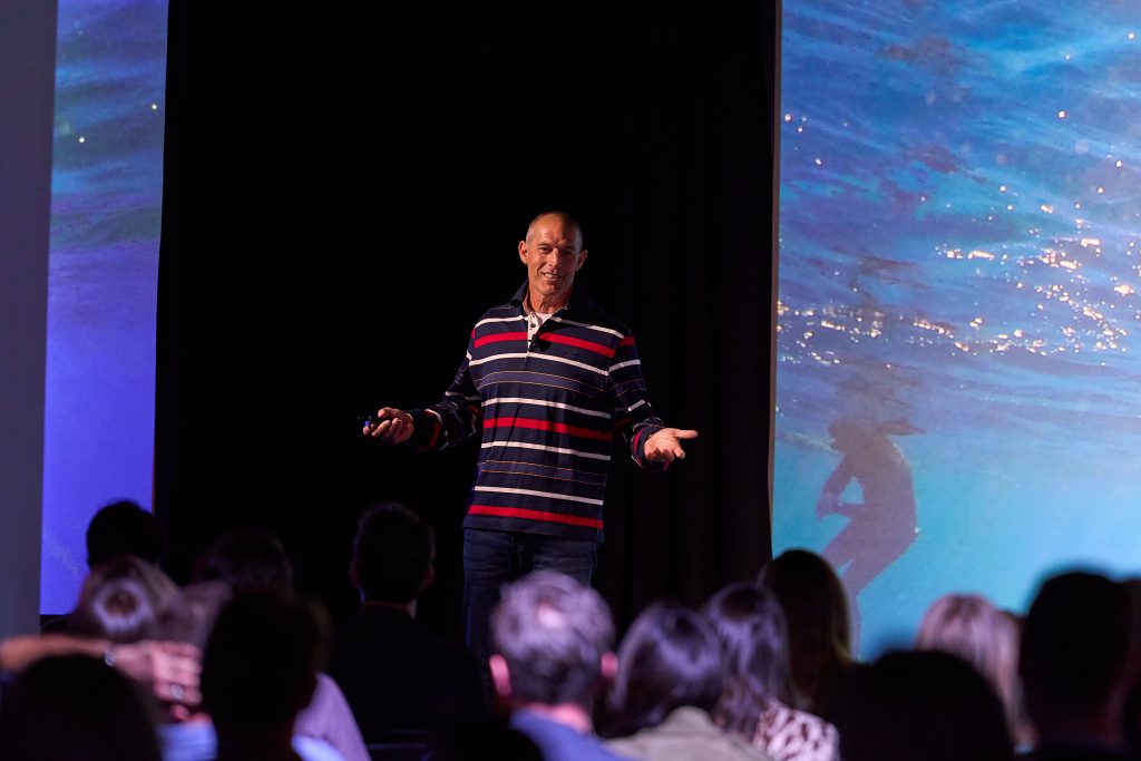 A man in a striped shirt stands on stage giving a presentation to an audience, with a large screen behind him displaying an underwater scene with a swimming figure.