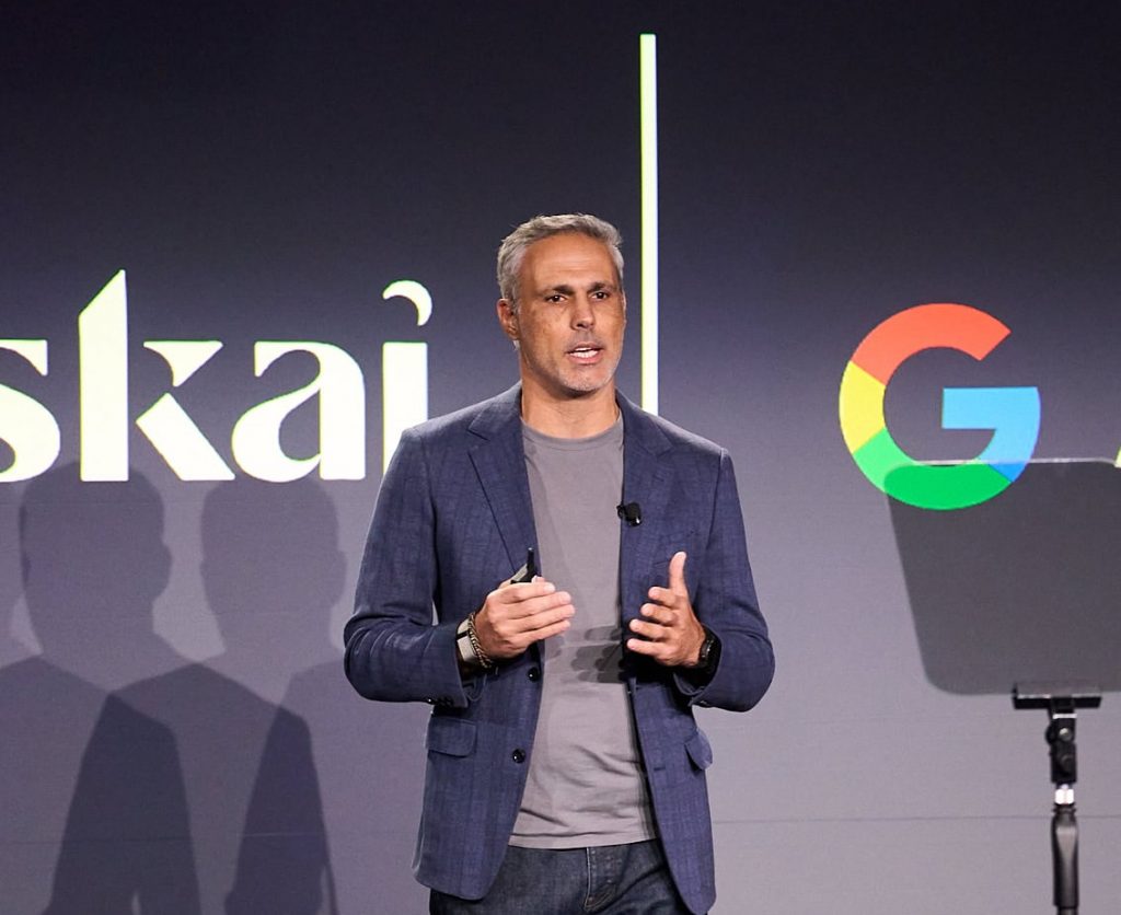 A man in a gray t-shirt and blue blazer speaks on stage with a microphone, standing in front of a screen displaying partial text and a colorful Google G logo.