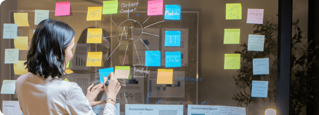 Dans un bureau, une femme écrit sur un tableau transparent recouvert de post-it colorés et de schémas, en réfléchissant à des idées liées à l'intelligence artificielle (IA).