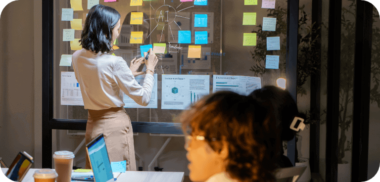 Une femme se tient devant un mur vitré recouvert de post-it colorés et de tableaux ; elle écrit ou classe des notes, tandis qu'au premier plan, deux personnes travaillent sur leurs ordinateurs portables, avec des tasses de café à proximité.