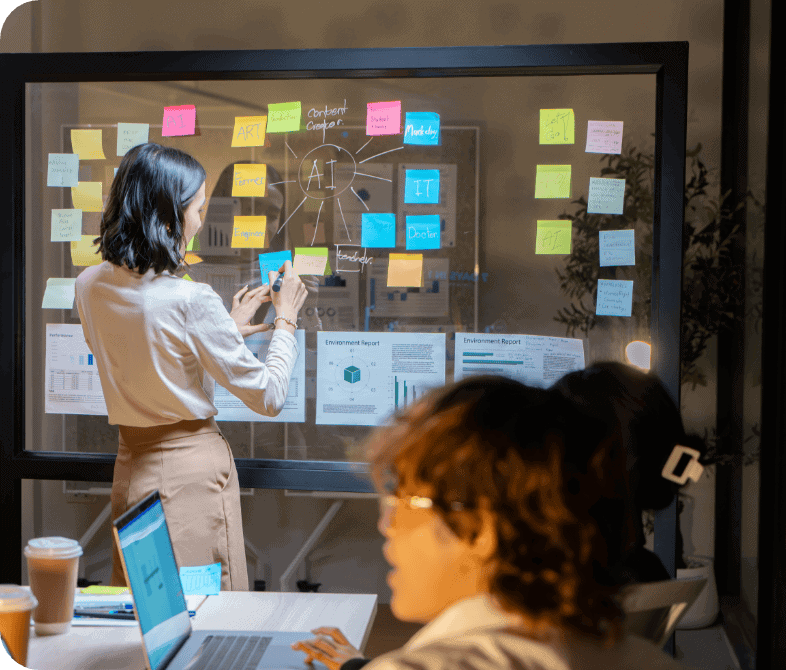 Dans un bureau au style moderne, une femme se tient debout devant un tableau en verre recouvert de post-it colorés et de schémas liés à l'IA, tandis qu'une autre personne travaille sur un ordinateur portable à une table voisine.