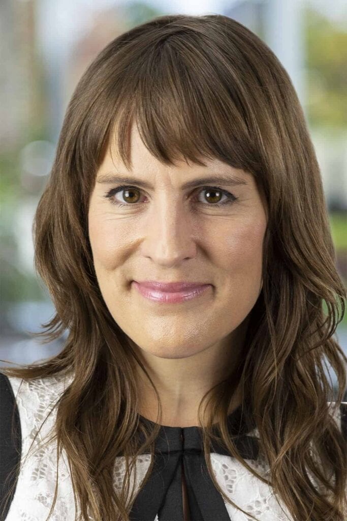 A woman with long brown hair and bangs, wearing a white top with a black collar, smiles softly at the camera with a blurred, greenish background behind her—capturing the warmth and approachability that reflects the importance of omnichannel connections in creating meaningful interactions.