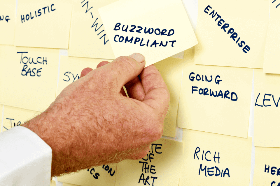 Une main appose un post-it sur lequel on peut lire « CONFORME AUX MOTS À LA MODE » sur un tableau recouvert d’autres notes rédigées dans un jargon d’entreprise, telles que « À L’AVENIR », « ENTREPRISE » et « l’importance de l’omnicanal ».