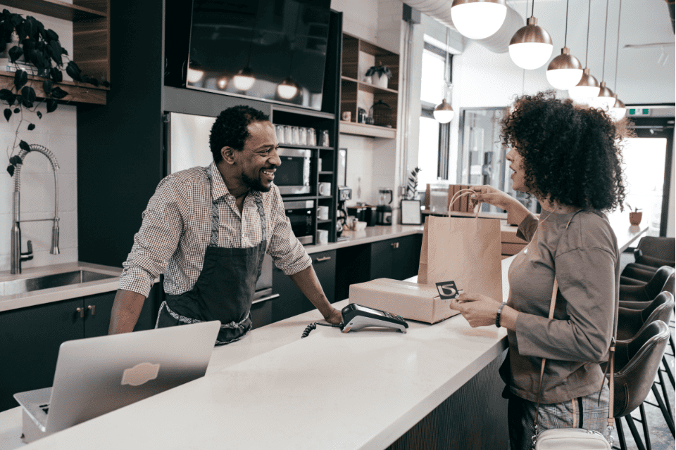 Ein Barista in einer Schürze lächelt einen Omnichannel-Kunden an, der ihm eine Karte über den Tresen reicht. Der Kunde erhält eine Papiertüte, im Hintergrund des Cafés sind Kaffeezubehör und Dekoration zu sehen.