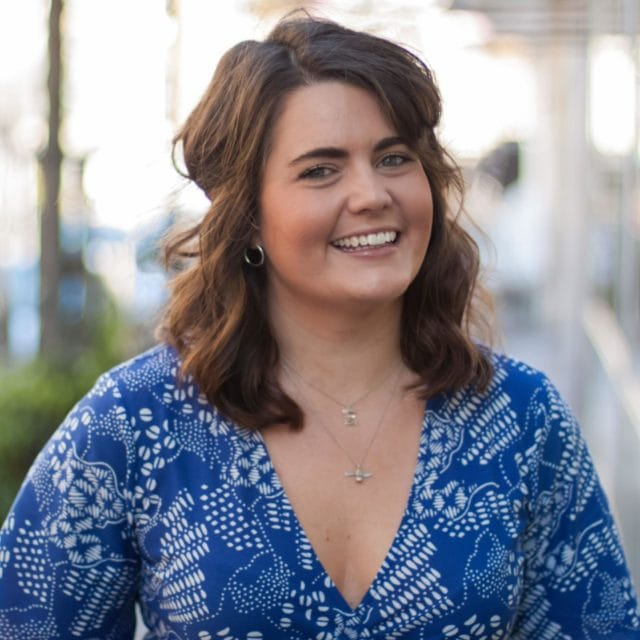 Woman with wavy brown hair wearing a blue patterned dress smiles outdoors, embodying the confidence of a modern omnichannel customer. She has silver hoop earrings and layered necklaces. The background is softly blurred, with hints of greenery and urban elements.