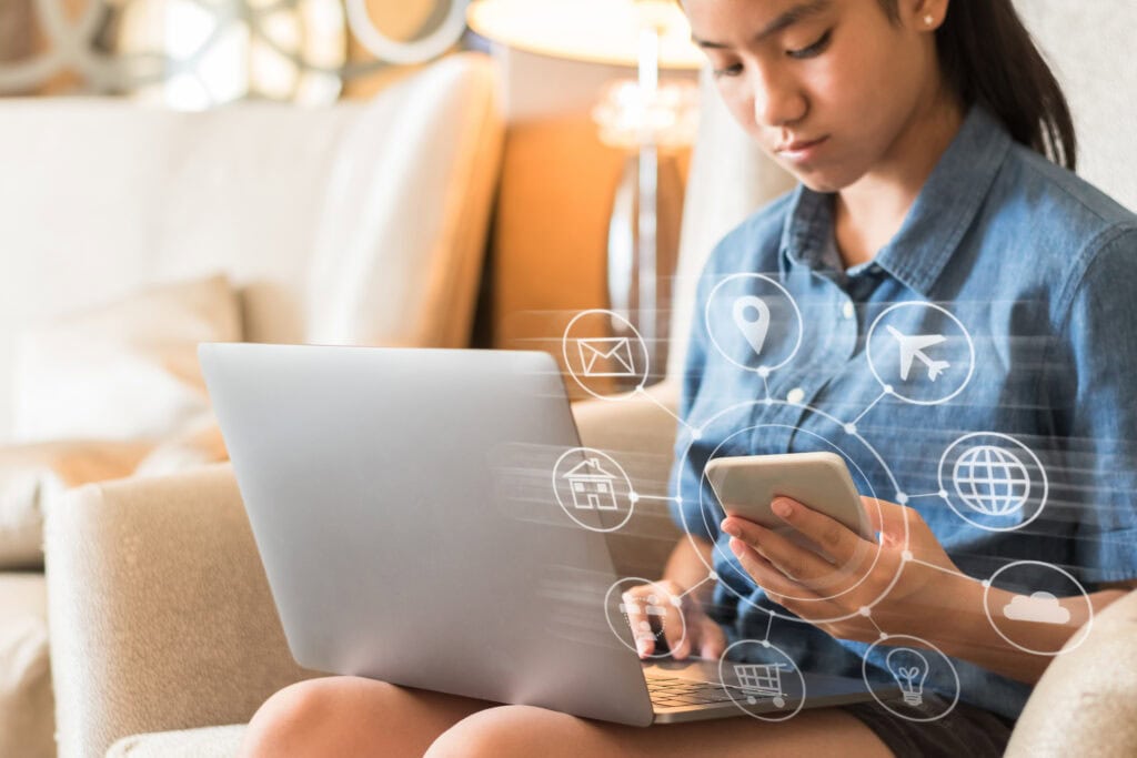 Eine junge Frau sitzt auf einem Sofa, arbeitet am Laptop und hält ein Smartphone in der Hand. Vor ihr erscheinen virtuell digitale Symbole für E-Mail, Standort, Flugzeug, Zuhause und Globus – was verdeutlicht, wie wichtig Omnichannel-Konnektivität in der heutigen digitalen Welt ist.