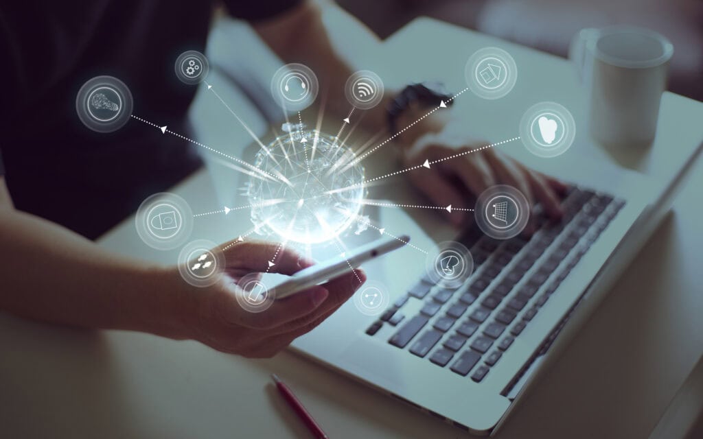 Eine Person, die ein Smartphone und einen Laptop benutzt, über den Geräten sind digitale Symbole und ein leuchtender Globus zu sehen, die für Vernetzung und Technologie stehen und die Bedeutung von Omnichannel-Erlebnissen unterstreichen. Im Hintergrund ist außerdem eine Kaffeetasse auf dem Tisch zu sehen.