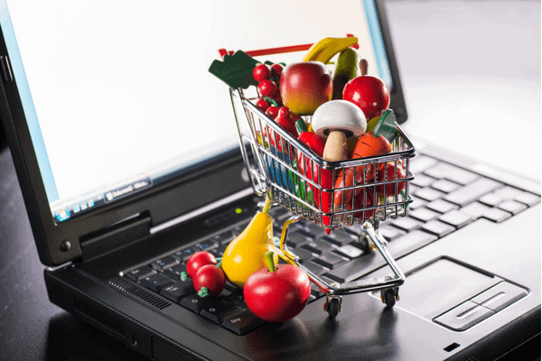 A small shopping cart filled with toy fruits and vegetables sits on the keyboard of an open laptop, illustrating the importance of omnichannel strategies in online grocery shopping.