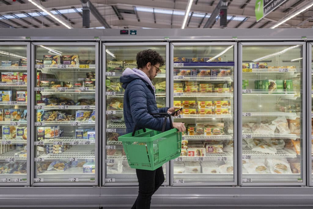 Eine Person mit einem grünen Einkaufskorb schaut auf ihr Handy, während sie vor einer Supermarkt-Tiefkühltruhe steht, die mit verschiedenen Tiefkühlprodukten gefüllt ist – das unterstreicht, wie wichtig Omnichannel-Erlebnisse beim modernen Lebensmitteleinkauf sind.