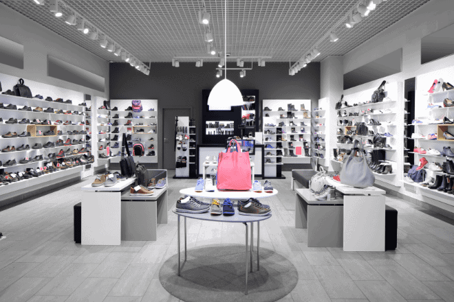 Une boutique de chaussures moderne, dont les murs sont bordés d'étagères blanches présentant divers modèles de chaussures et d'accessoires. Mettant l'accent sur l'importance de l'omnicanal, cet espace allie harmonieusement la découverte en magasin aux avantages du commerce en ligne. Au centre, des tables exposent d'autres modèles de chaussures et de sacs à main sous un éclairage vif, tandis que le sol est recouvert de carrelage gris clair.