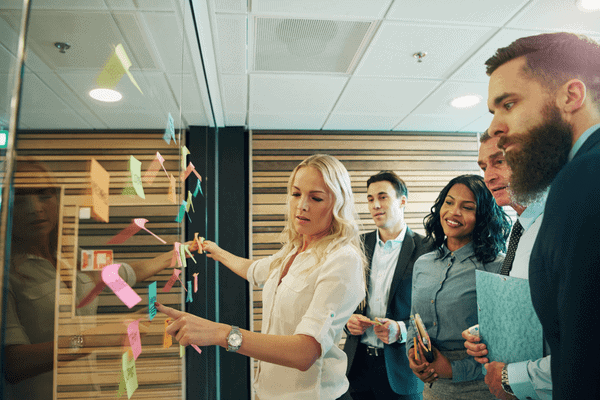 Five professionals stand by a glass wall covered in colorful sticky notes, as one woman points to the notes while others watch and listen attentively, suggesting a collaborative meeting or brainstorming session focused on the importance of omnichannel strategies.