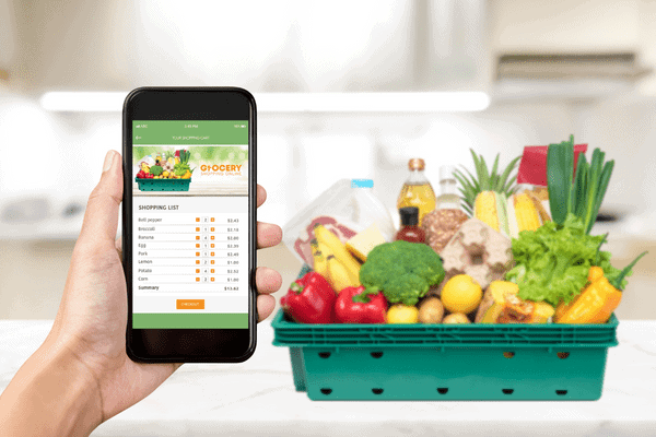 Eine Hand hält ein Smartphone, auf dem eine App für den Lebensmitteleinkauf zu sehen ist, was die Bedeutung des Omnichannel-Shoppings unterstreicht; im Hintergrund steht ein Korb mit frischem Gemüse, Obst und Lebensmitteln auf einer Küchentheke.