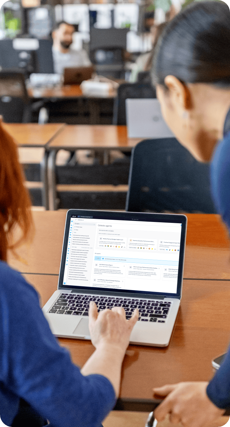 Two people look at a laptop screen displaying data dashboards with graphs and charts, discussing insights from AI marketing consulting services, seated at a desk in an office with other people working in the background.
