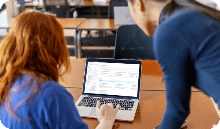 Two people look at a laptop screen displaying data charts and graphs in a classroom or office setting with wooden tables and chairs in the background, as they discuss strategies provided by AI marketing consulting services.